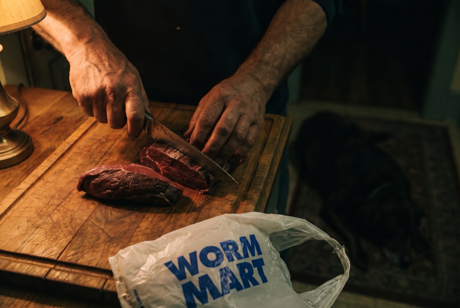 A man cuts raw steak in a dim kitchen, a Worm Mart grocery bag in the foreground, a dog in the shadows